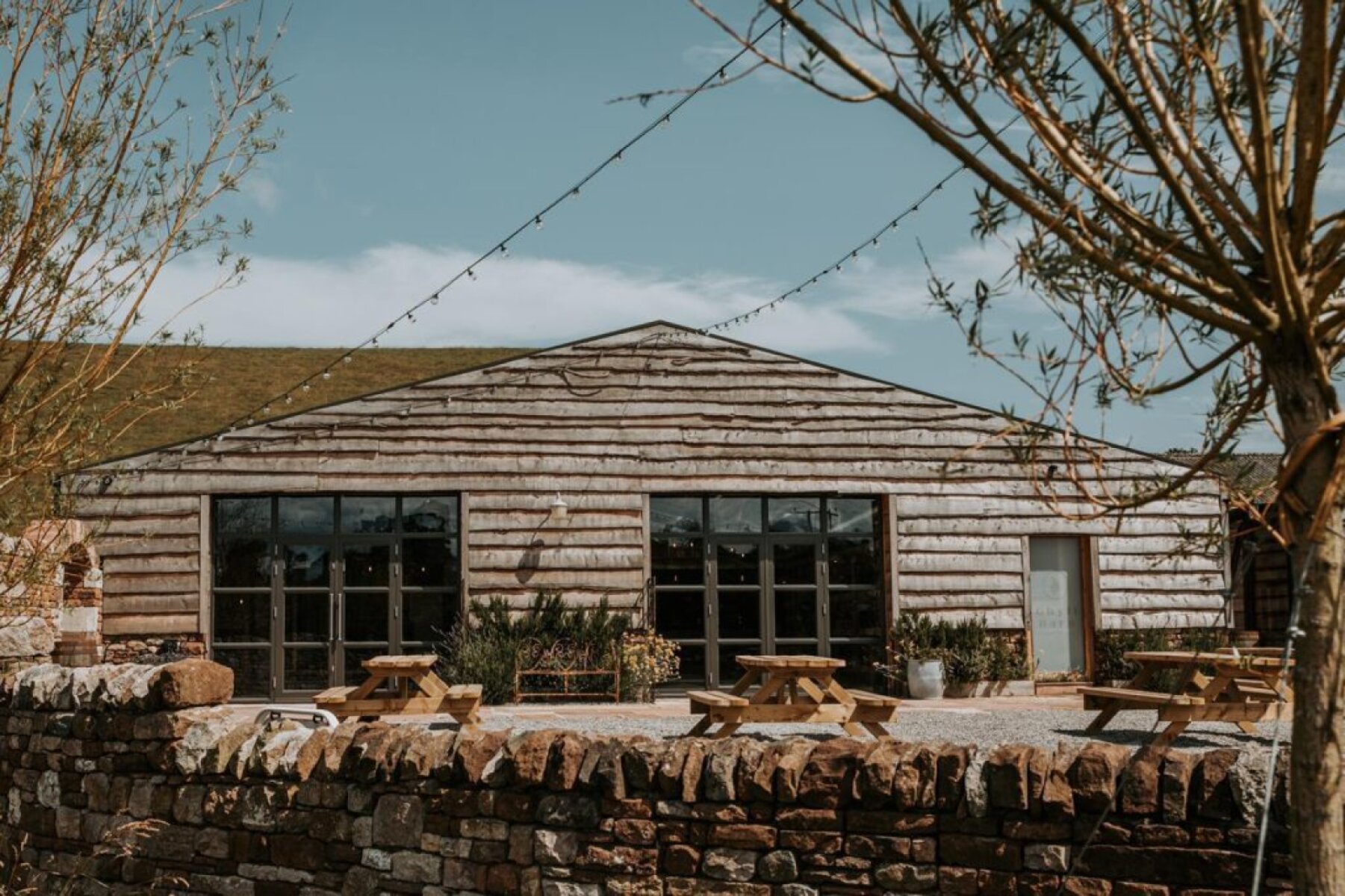 Charming rustic barn venue with large windows, surrounded by greenery and picnic tables, perfect for a relaxed outdoor wedding.