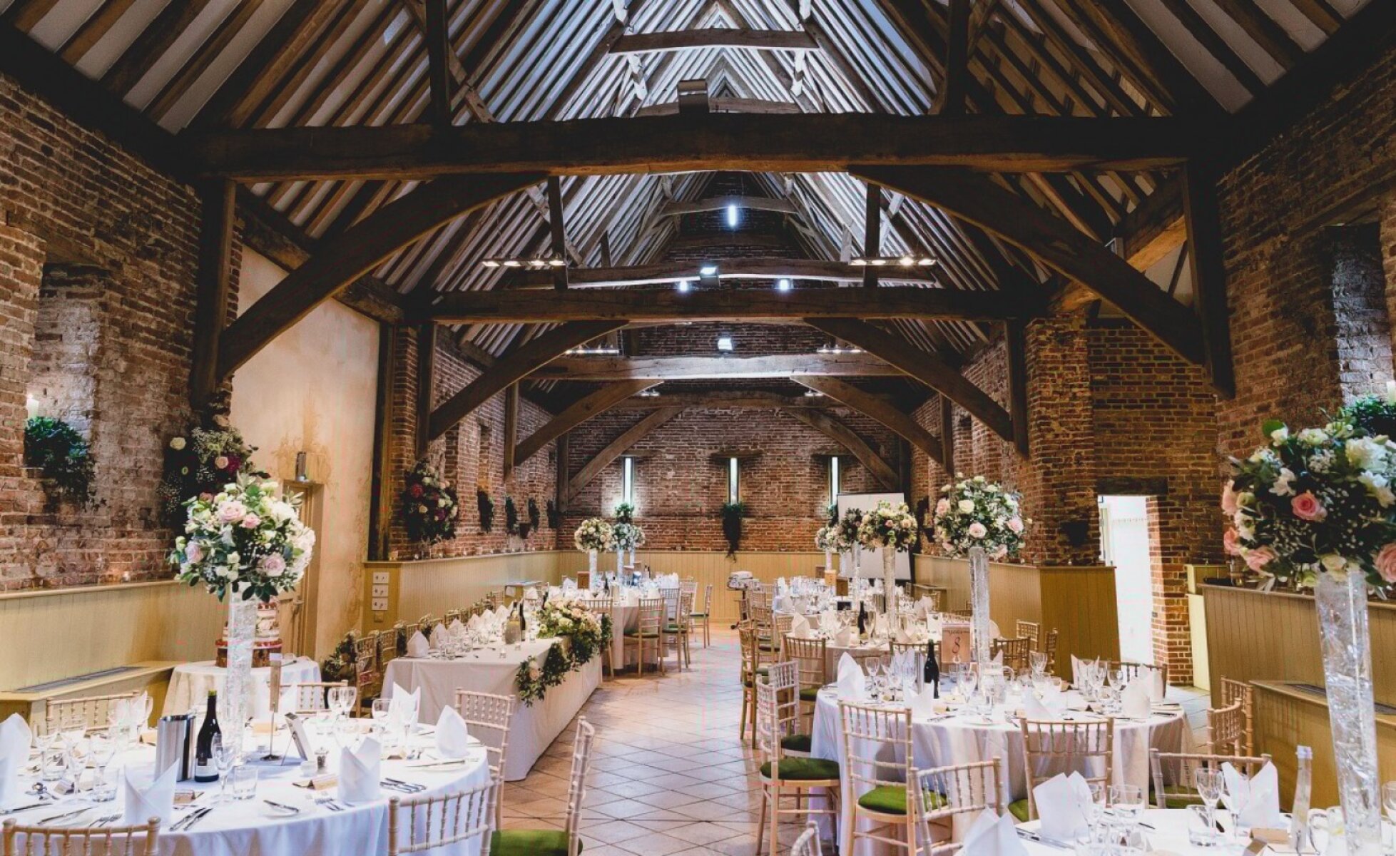 A rustic barn interior with high, wooden beams, elegant tables adorned with flowers, and soft lighting creating a romantic atmosphere.