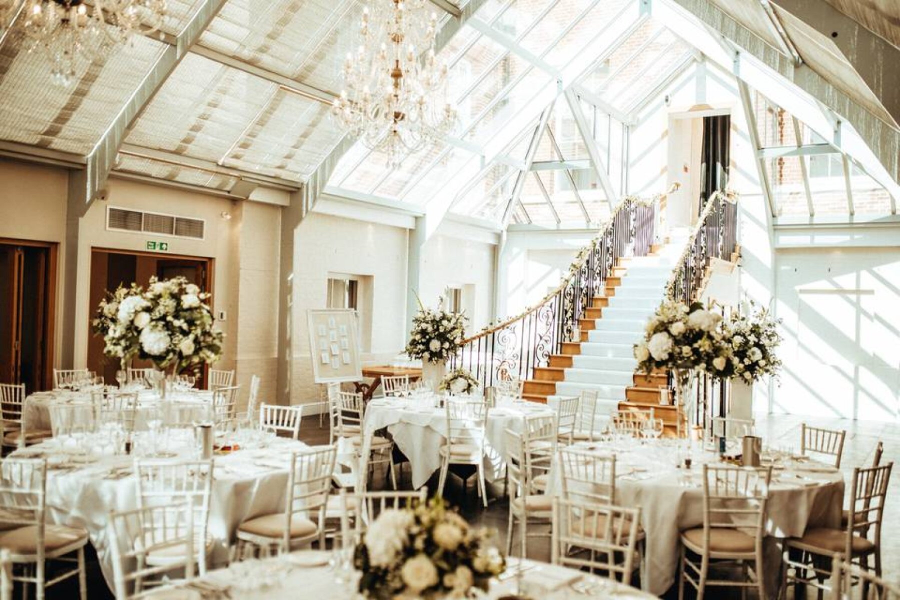 Bright and airy wedding venue featuring elegant round tables, floral centerpieces, and a stunning staircase under a glass roof.