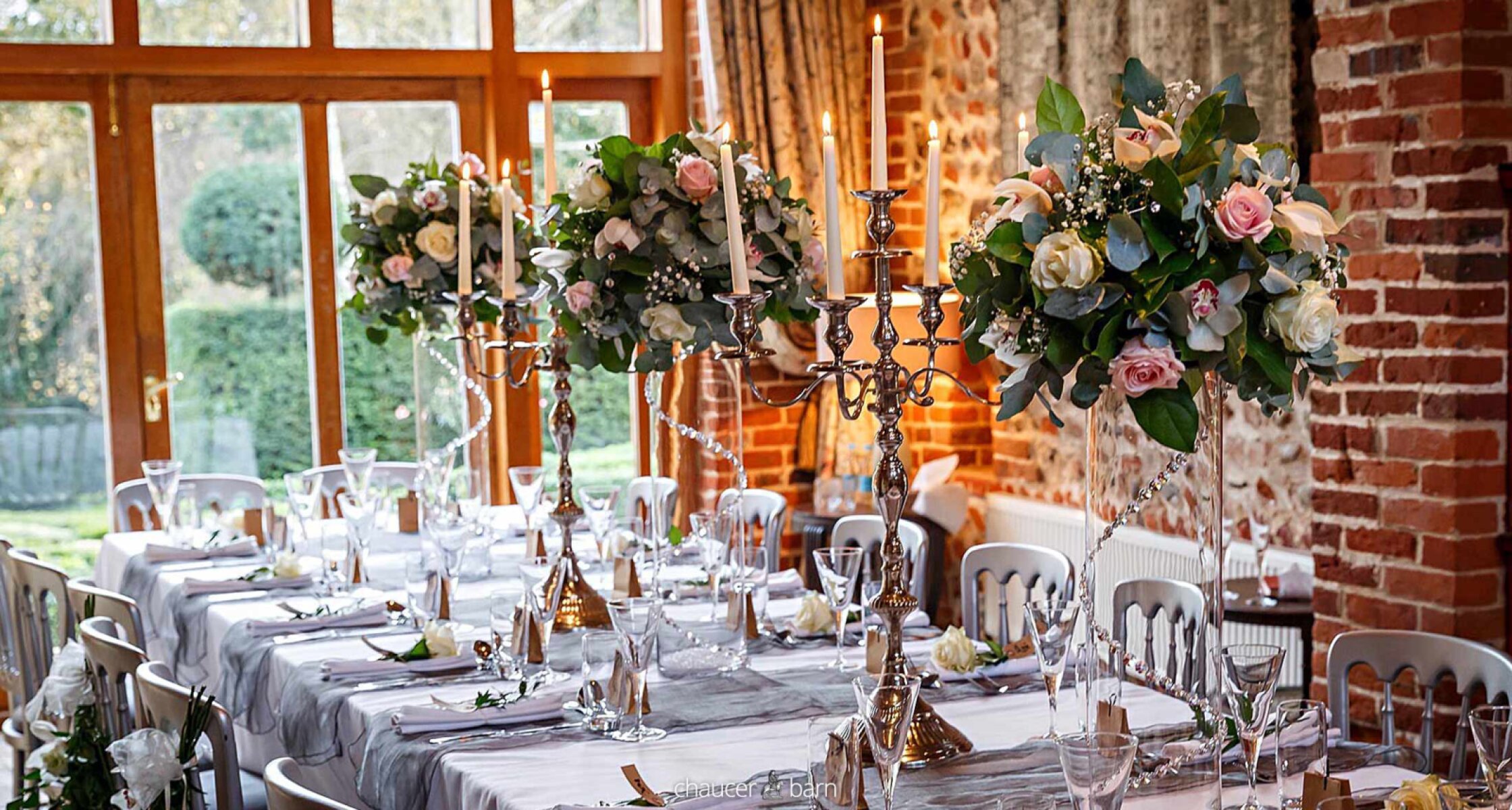 Elegant reception table set with floral centerpieces, candle holders, and soft gray linens, featuring large windows and natural light.
