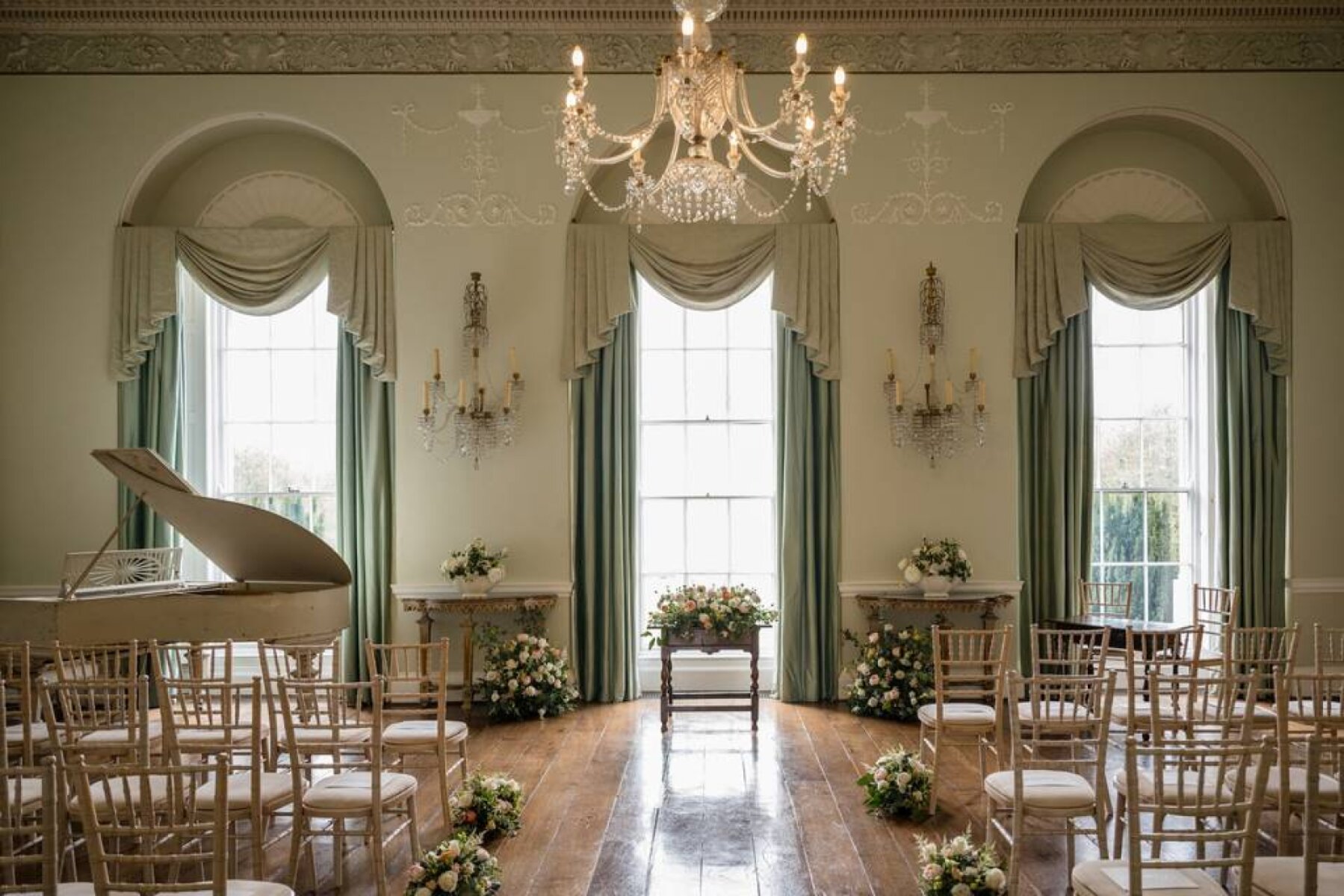Elegant ceremony space featuring large windows, chandelier, hardwood floors, and beautifully arranged floral accents. Chic and stylish atmosphere.