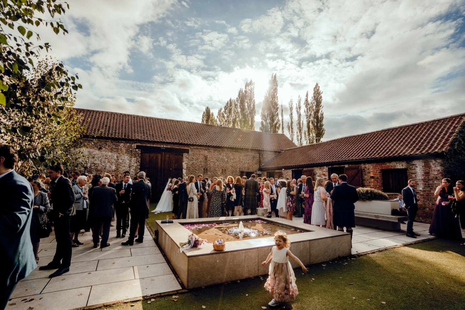 Charming outdoor wedding scene with guests mingling around a central stone fountain, framed by rustic buildings and wooded backdrop.