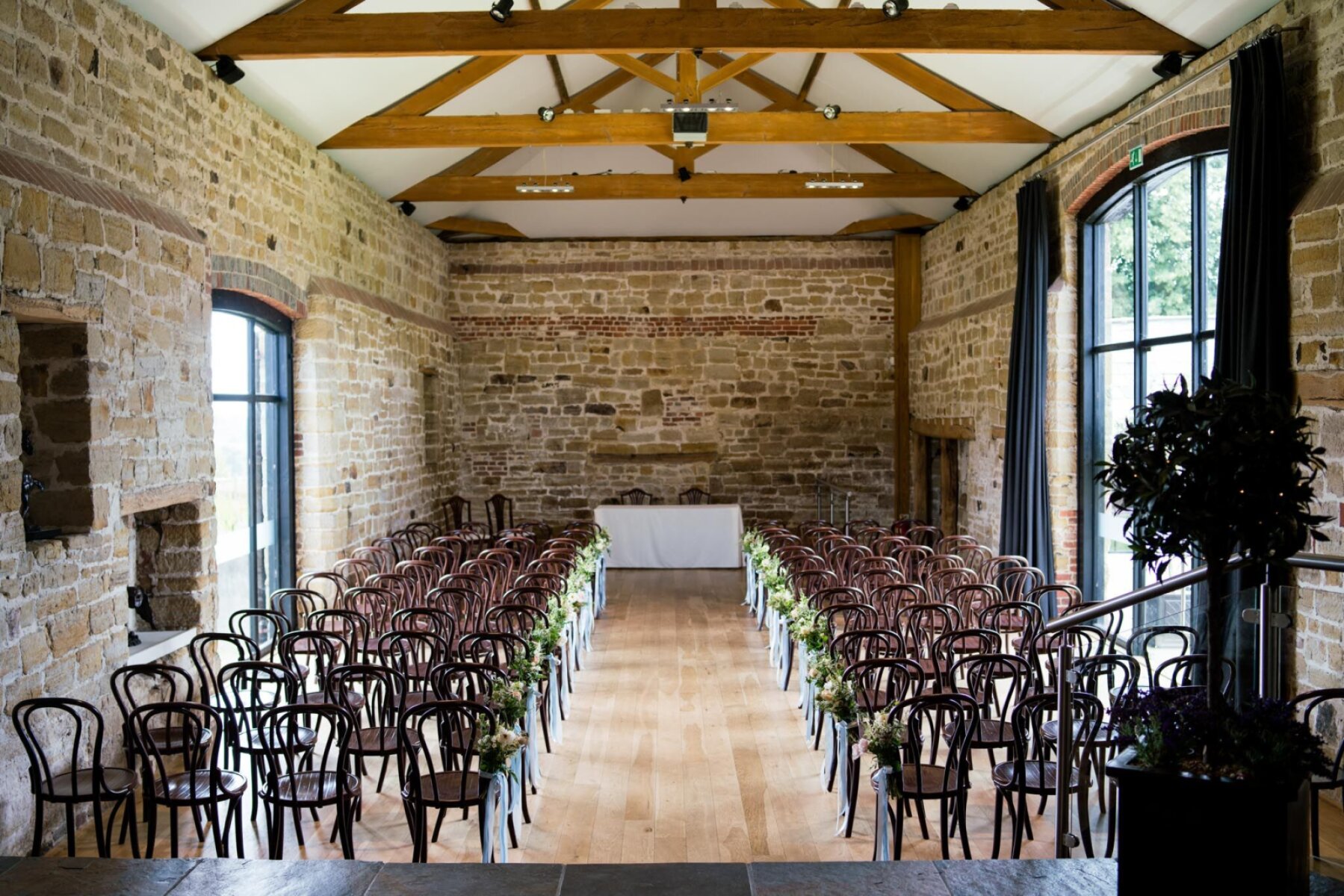 Rustic ceremony space with exposed stone walls, wooden beams, and rows of elegant chairs adorned with greenery. Bright, inviting atmosphere.