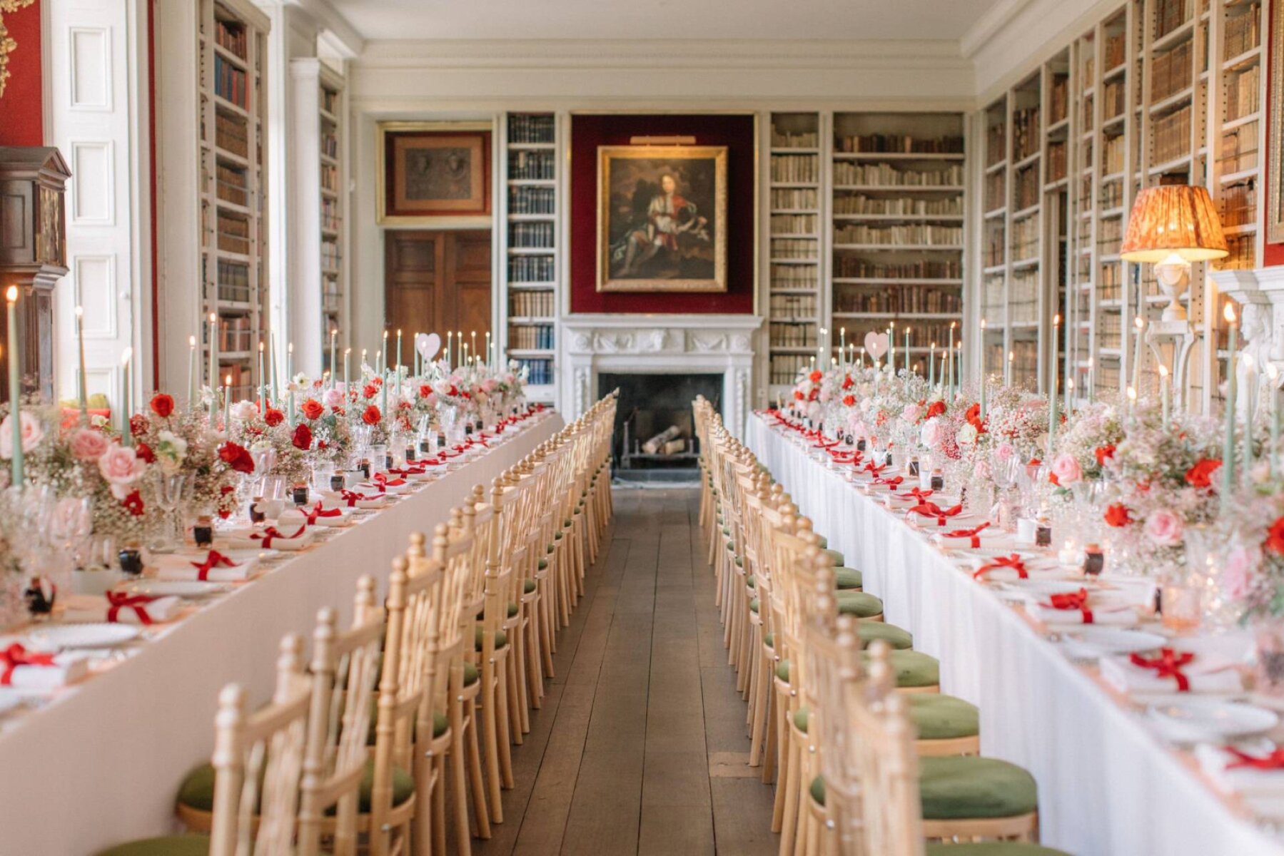 Elegant dining setup in a library with long tables adorned with pink flowers, candles, and golden chairs, creating a romantic ambiance.