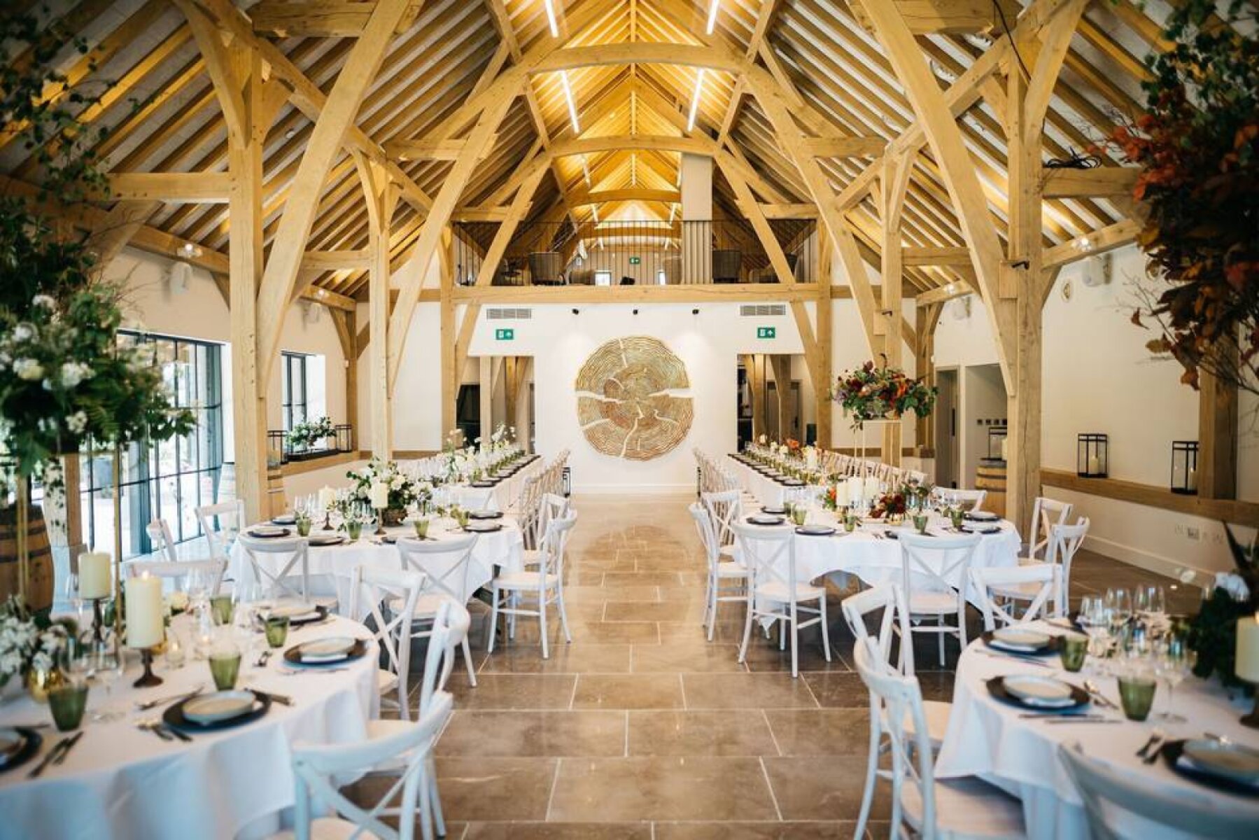 Bright, spacious barn venue featuring wooden beams, elegant tables set for a wedding, and lush greenery accents throughout.