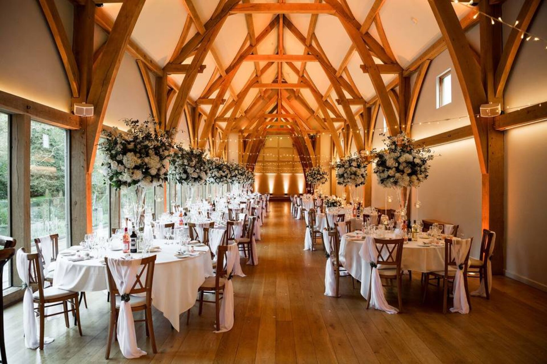 Elegant reception hall featuring tall wooden beams, spacious tables adorned with floral centerpieces, and warm ambient lighting.