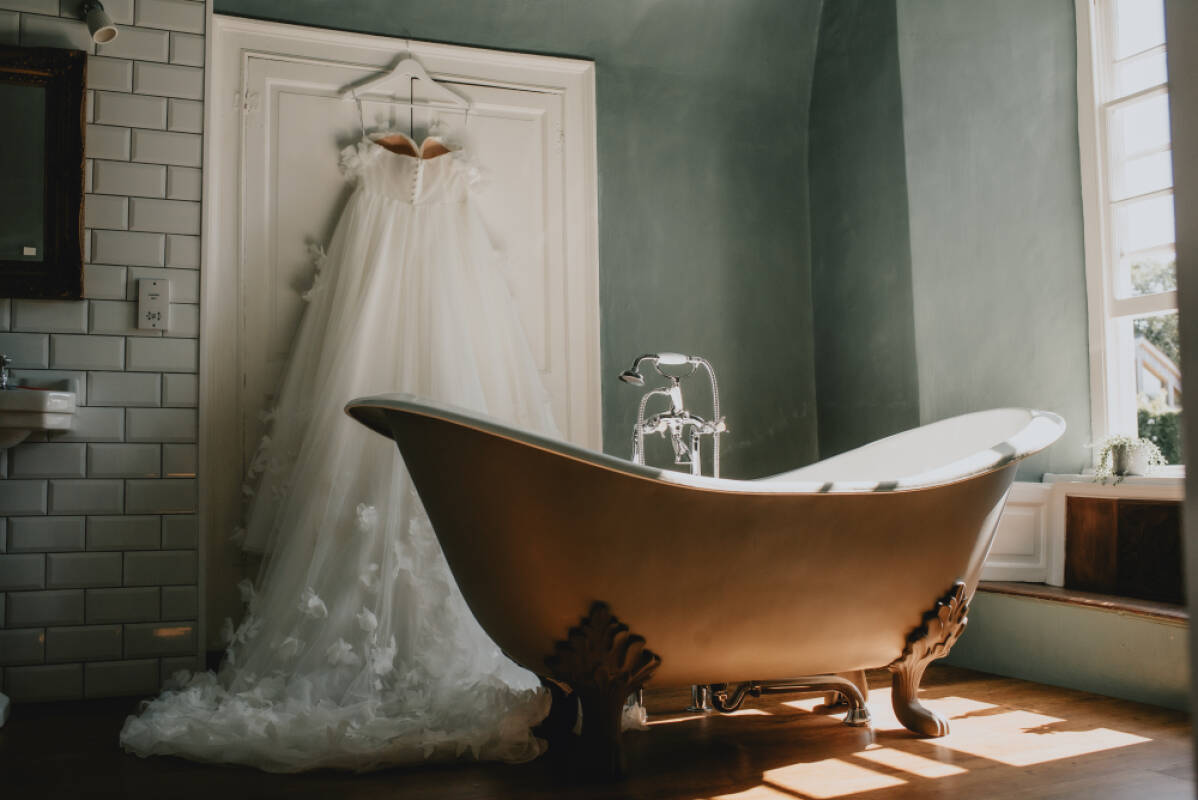A vintage clawfoot bathtub sits beneath a sunny window, showcasing a stunning wedding dress elegantly hanging on the wall.