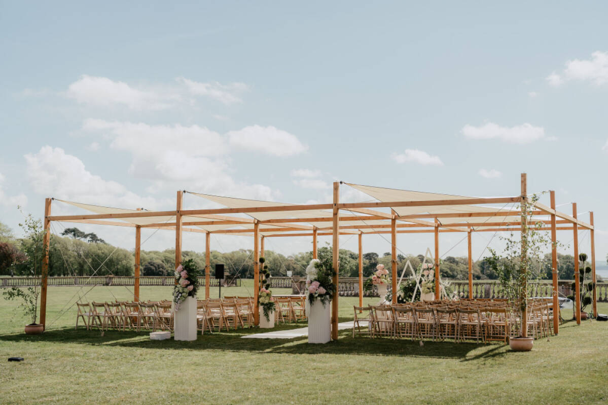 An elegant outdoor ceremony space featuring a wooden pergola, draped fabric, and stylish seating surrounded by greenery.