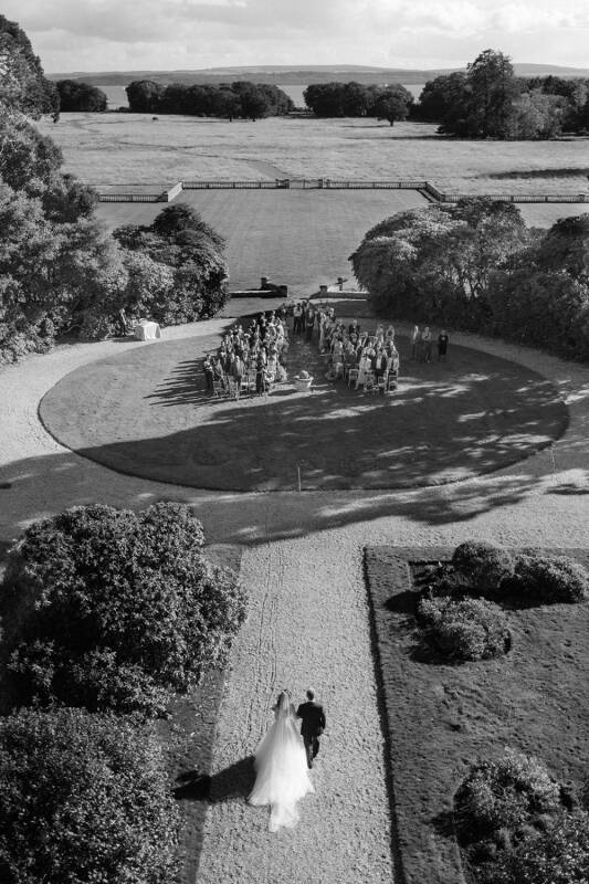 A couple walks towards a circular ceremony space surrounded by greenery, with serene water and a scenic landscape in the background.