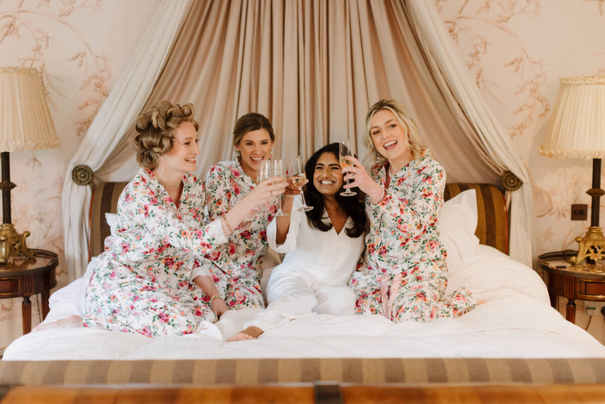 Bridesmaids in floral robes celebrate with champagne on a luxurious bed, featuring elegant decor and soft, neutral tones.